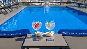 Vista de la piscina con dos vasos de una bebida transparente, un bol de aperitivos y un paisaje urbano al fondo.