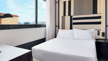 A hotel room with a bed, a bedside table, a nightstand, and a view of the cityscape through a large window.