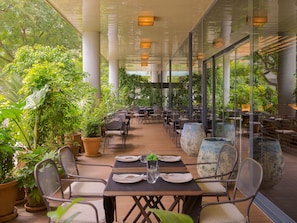 An outdoor dining area with tables and chairs, surrounded by potted plants and large windows.