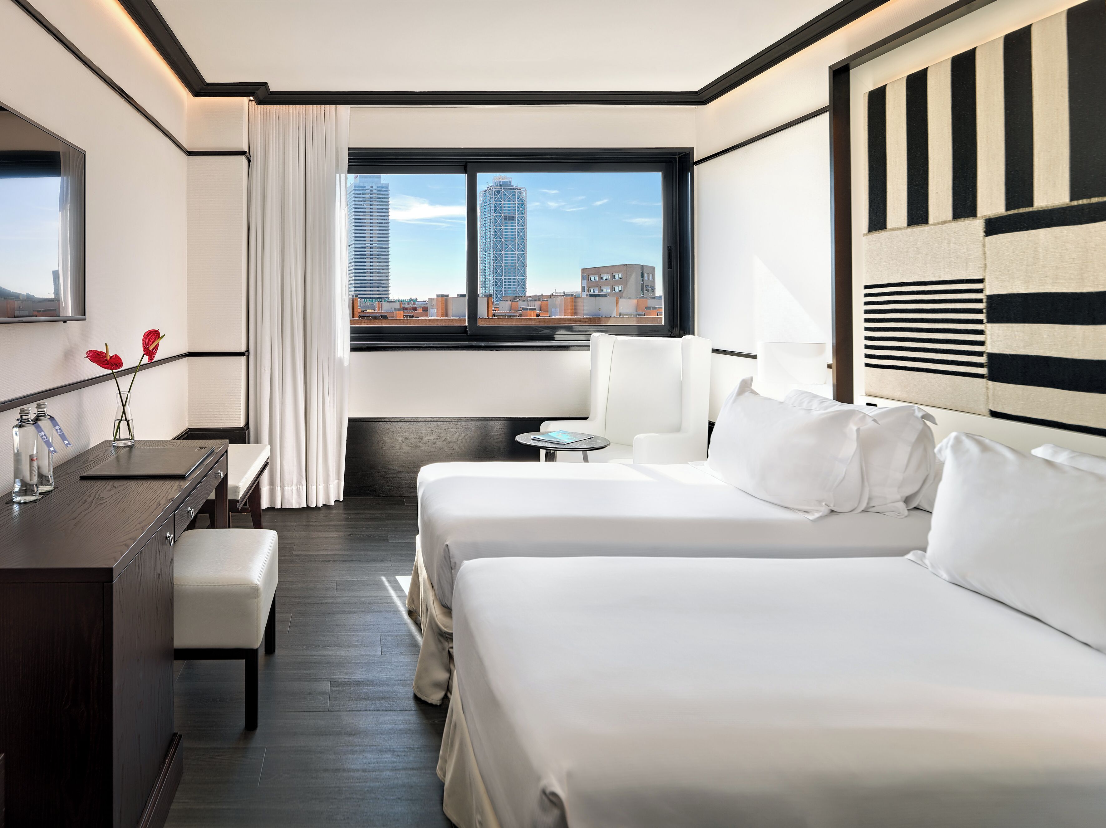A hotel room with two beds, a desk, a chair, a small table, and a view of the cityscape.