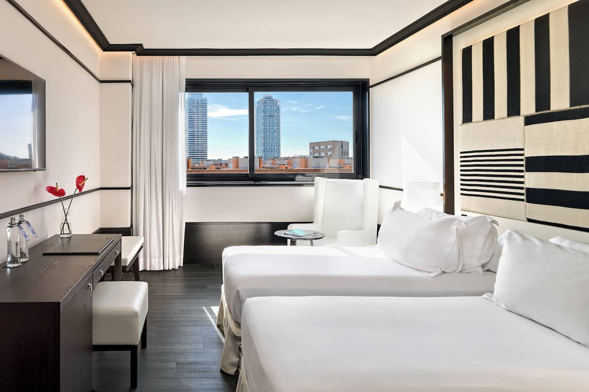 Ein Hotelzimmer mit zwei Betten, einem Schreibtisch, einem Sessel, einem kleinen Tisch und Blick auf die Stadt.