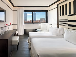 A hotel room with two beds, a desk, a chair, a small table, and a view of the cityscape.