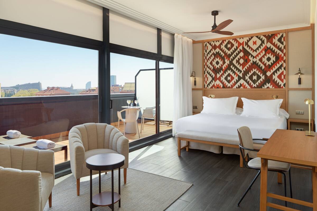 Ein Hotelzimmer mit einem großen Bett, einem Deckenventilator, einem Esstisch und einem Balkon mit Blick auf die Stadt.