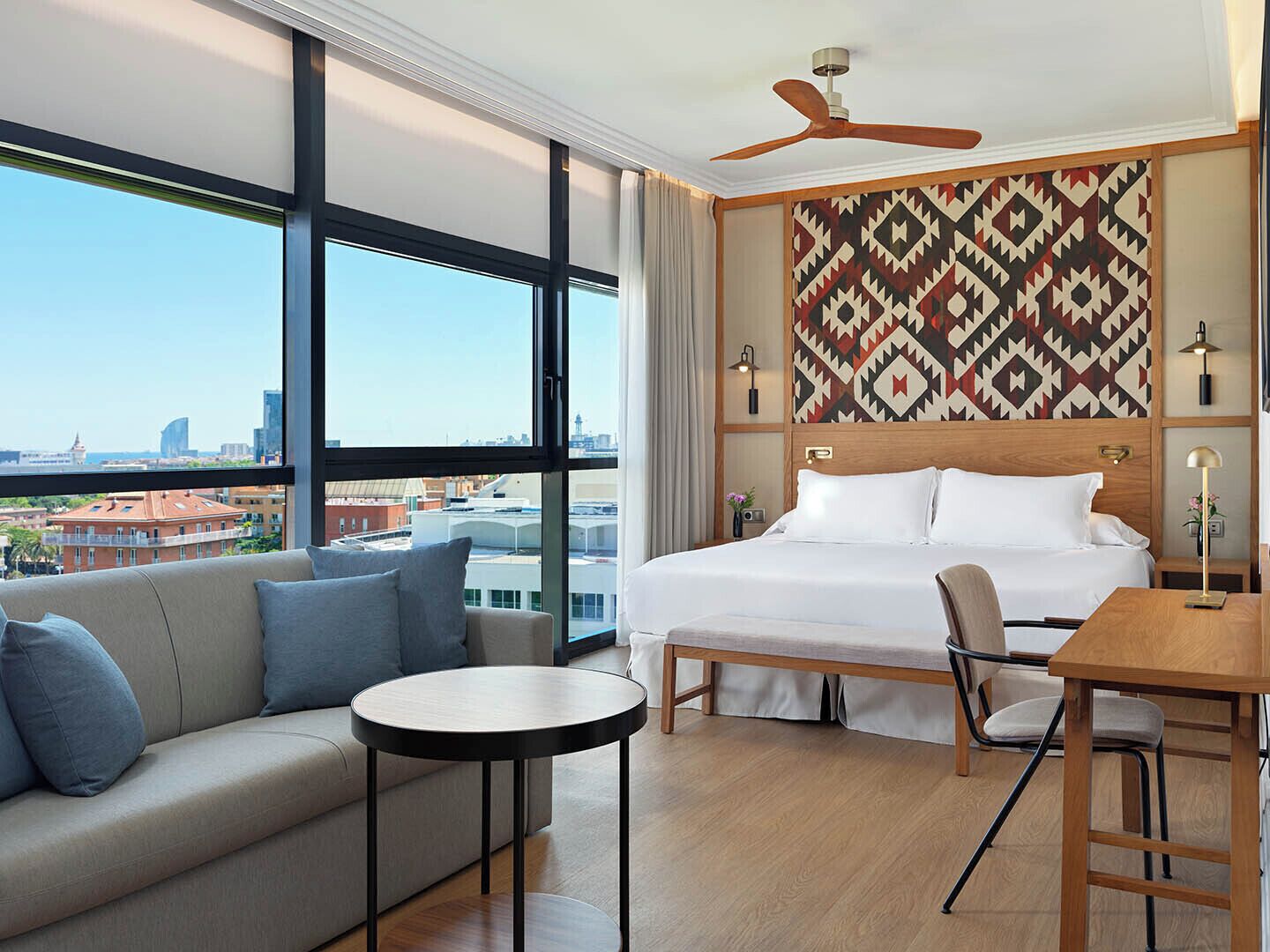 A hotel room with a large bed, a sofa, a dining table, and a view of the cityscape through large windows.