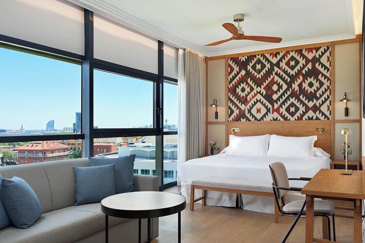 Ein Hotelzimmer mit einem großen Bett, einer Couch, einem Esstisch und Blick auf die Stadt durch große Fenster.