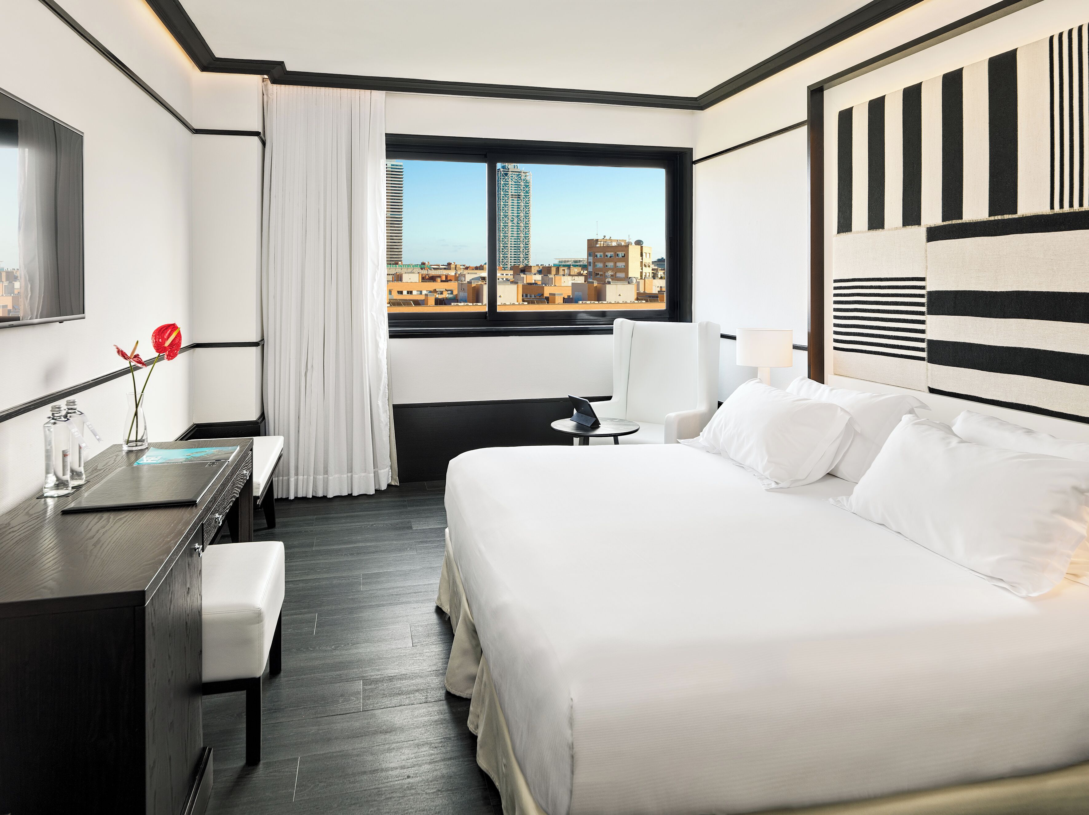 A hotel room with a large bed, a desk with a laptop, a chair, and a view of the cityscape through the window.