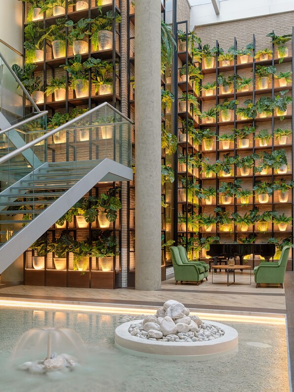 A modern indoor space with a staircase, a piano, green chairs, and a water feature.