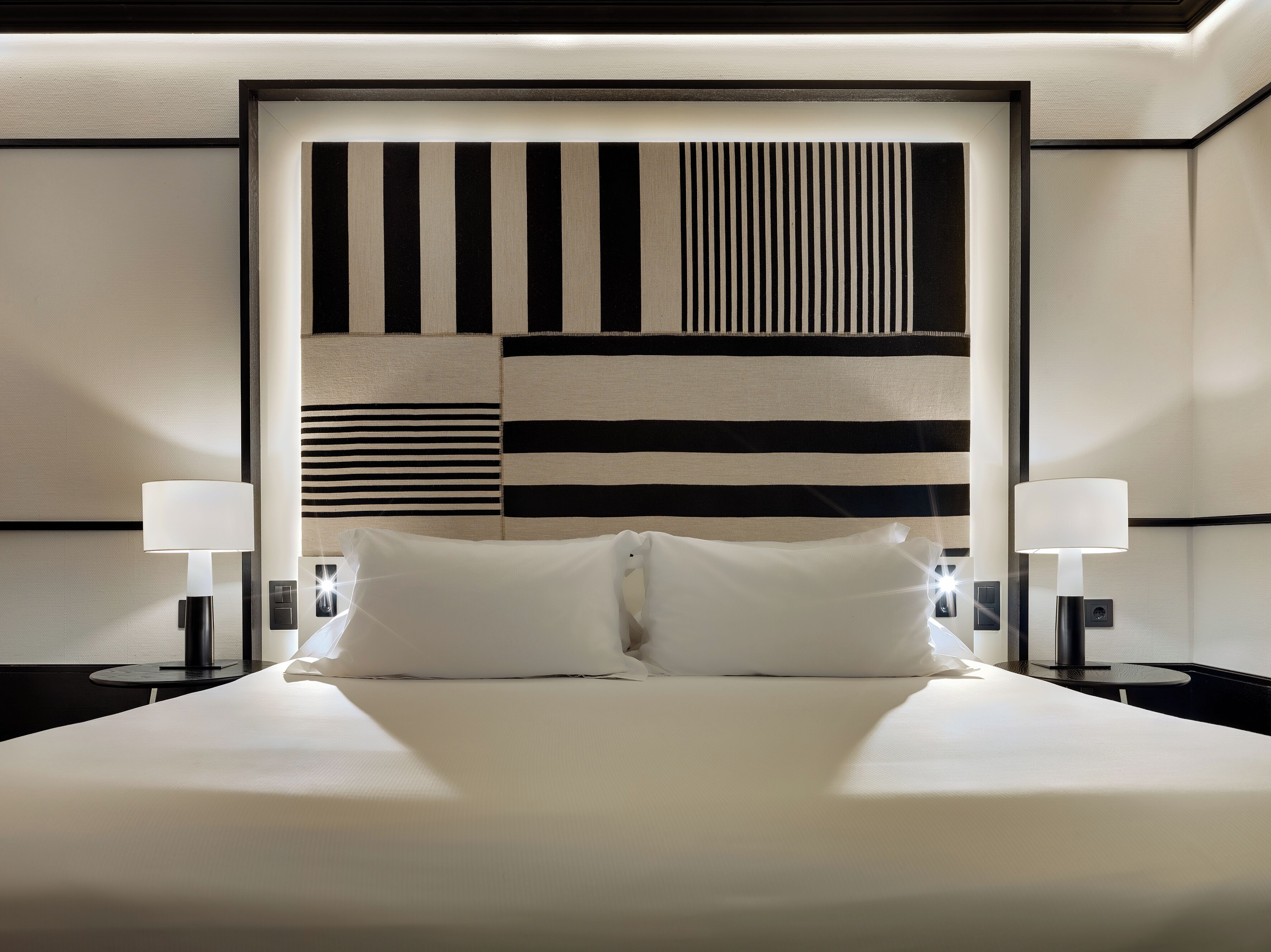 A neatly made bed with white linens, two bedside lamps, and a headboard with a geometric pattern.
