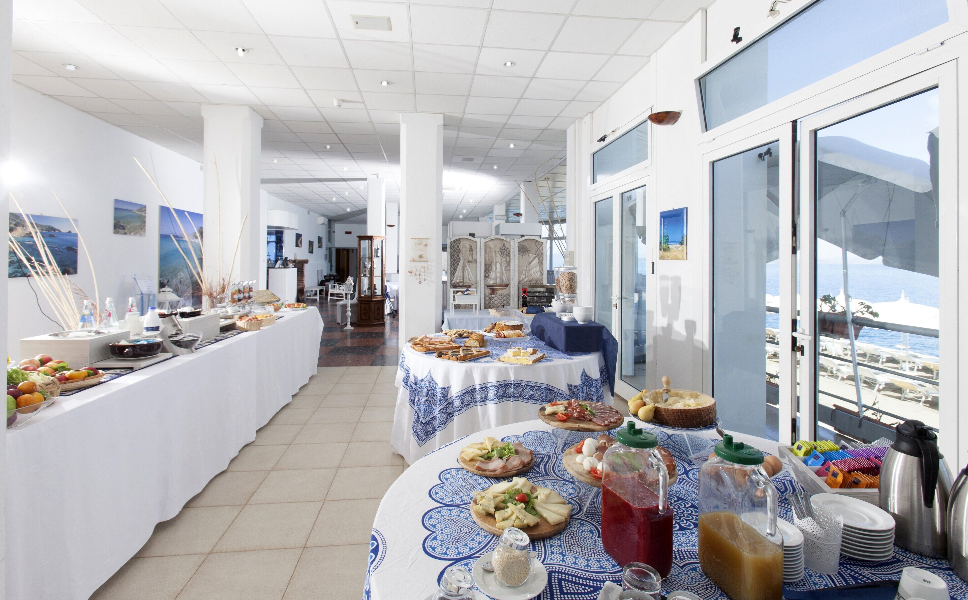 A buffet setup with various food items and beverages, a view of the sea through large windows, and a modern interior design.