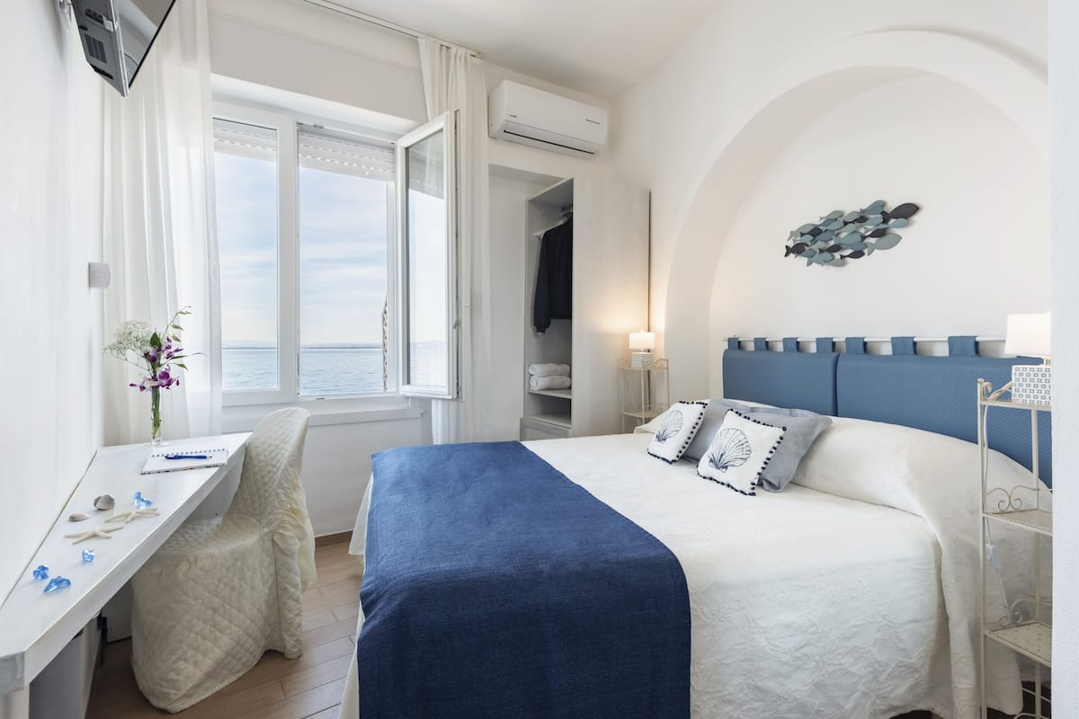 A bedroom with a bed, a desk, a chair, a window with curtains, an air conditioning unit, and a wall-mounted shelf.