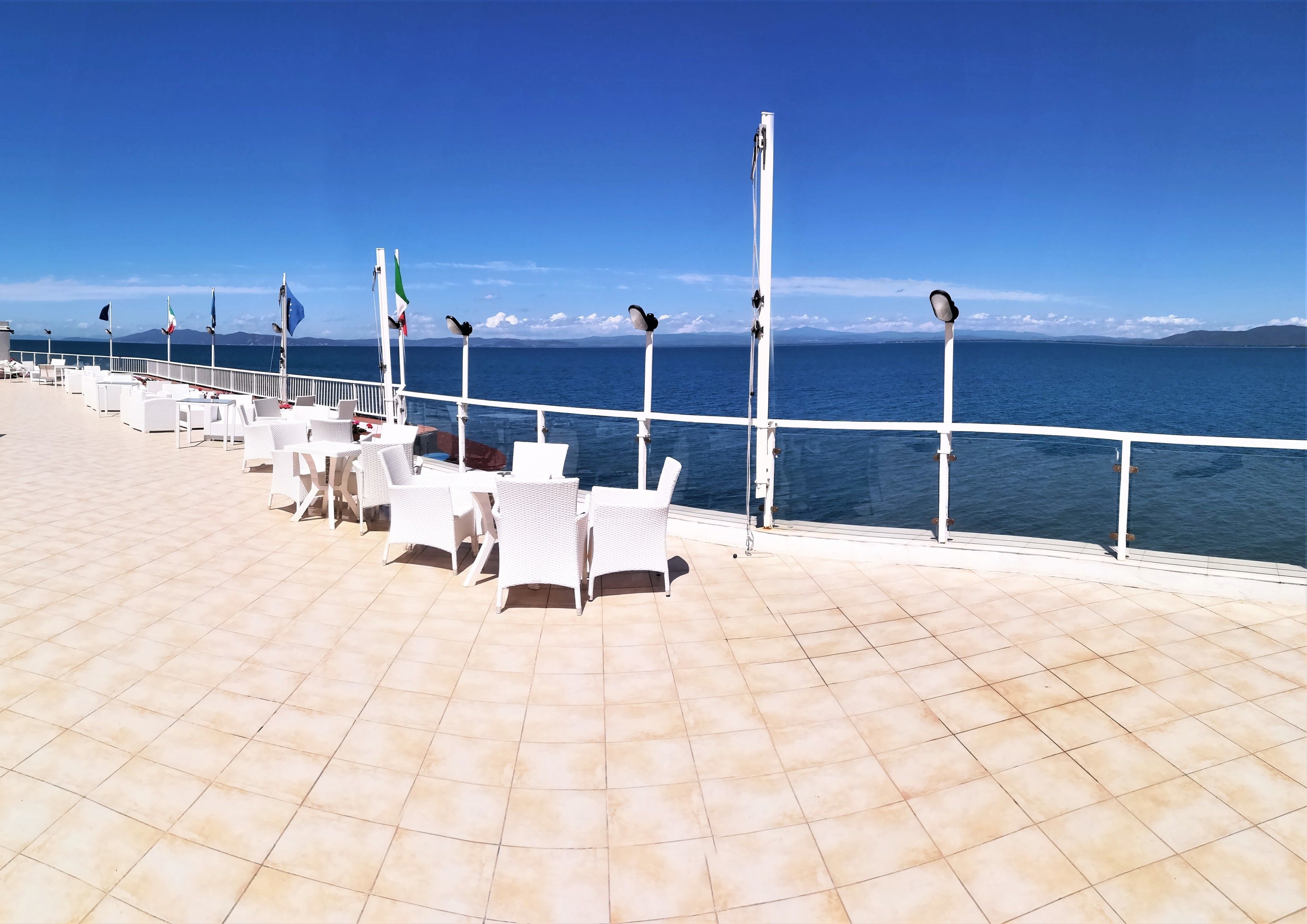 A sunlit terrace with white wicker chairs and tables, overlooking a calm sea and distant mountains.