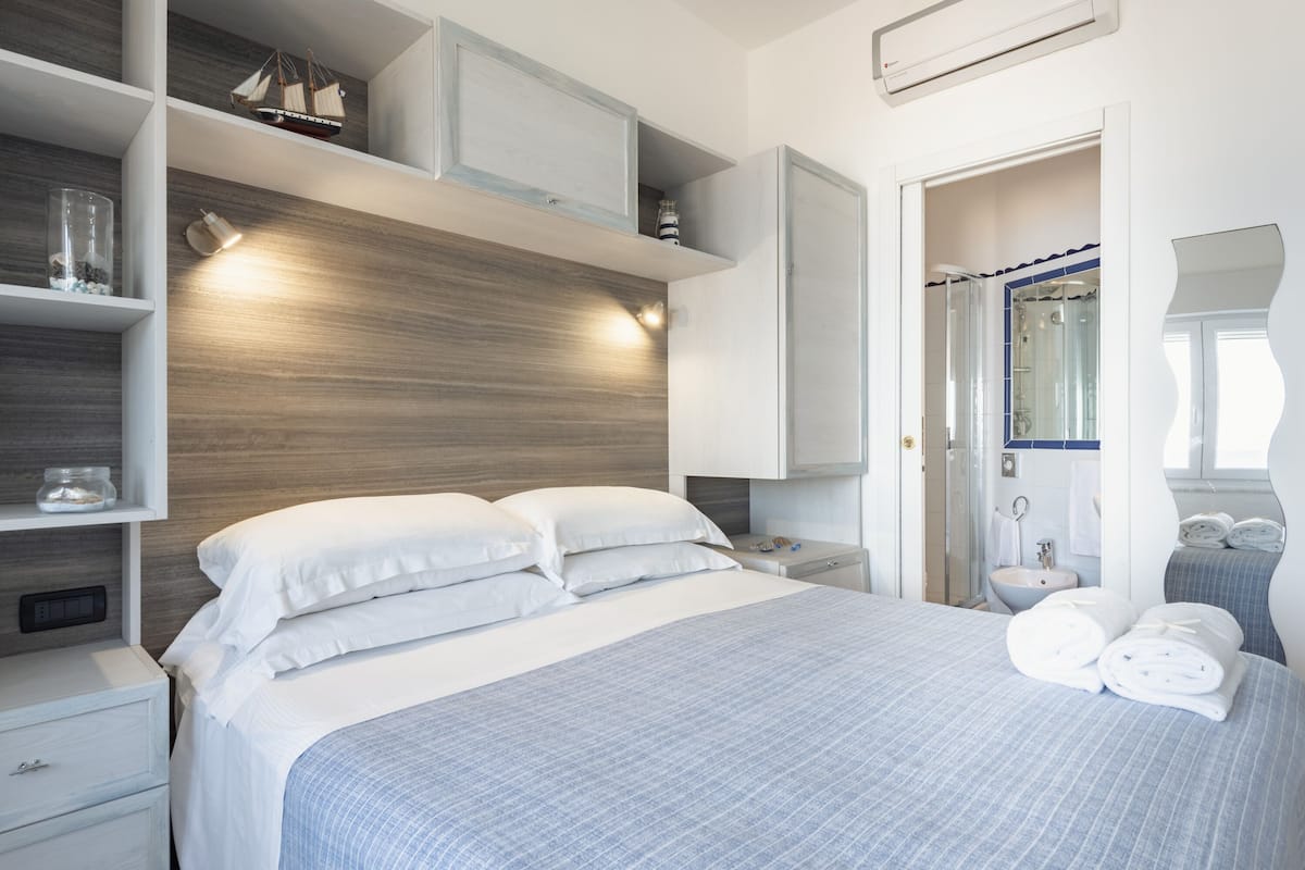 A modern hotel room with a large bed, a nightstand, a built-in shelf with decorative items, a bathroom with a sink and mirror, and a walk-in closet.