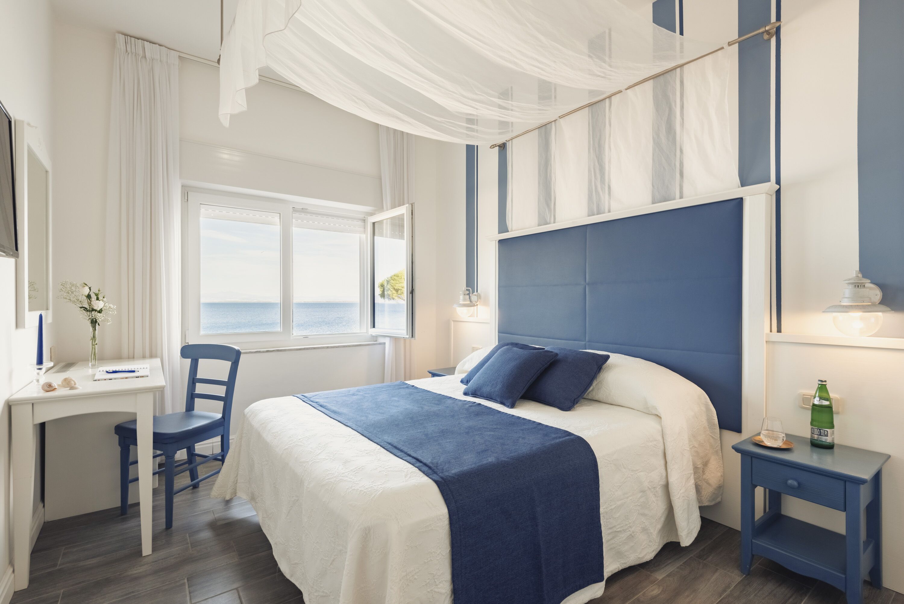 A bedroom with a bed, a desk, and a chair. There is a window with curtains, a nightstand, and a lamp.