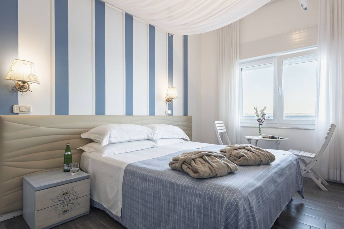 A neatly made bed with a blue and white striped headboard, a bedside table with a lamp, and a chair by the window.