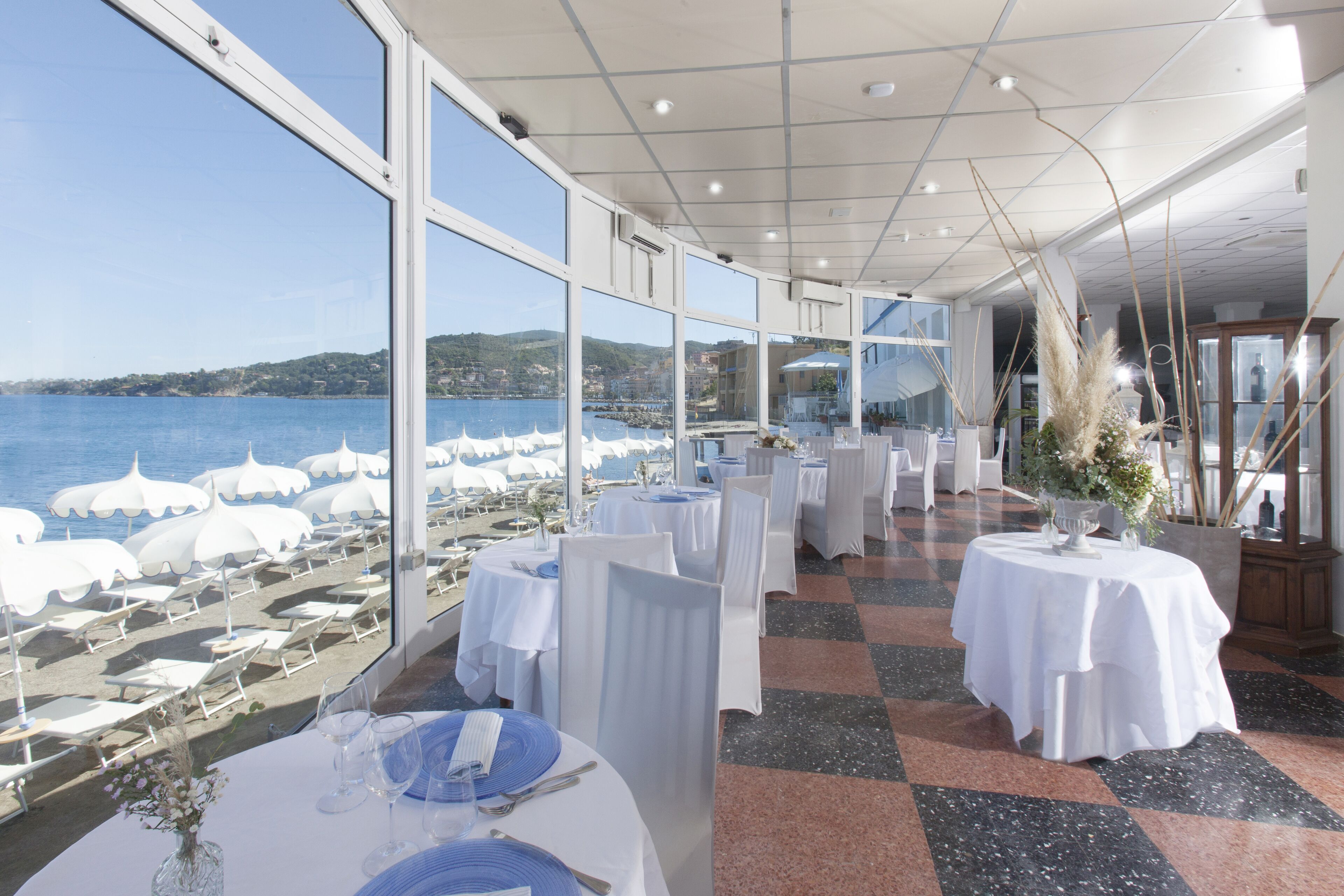 A restaurant with tables set for dining, large windows with a view of the sea and hills, and a checkered floor.