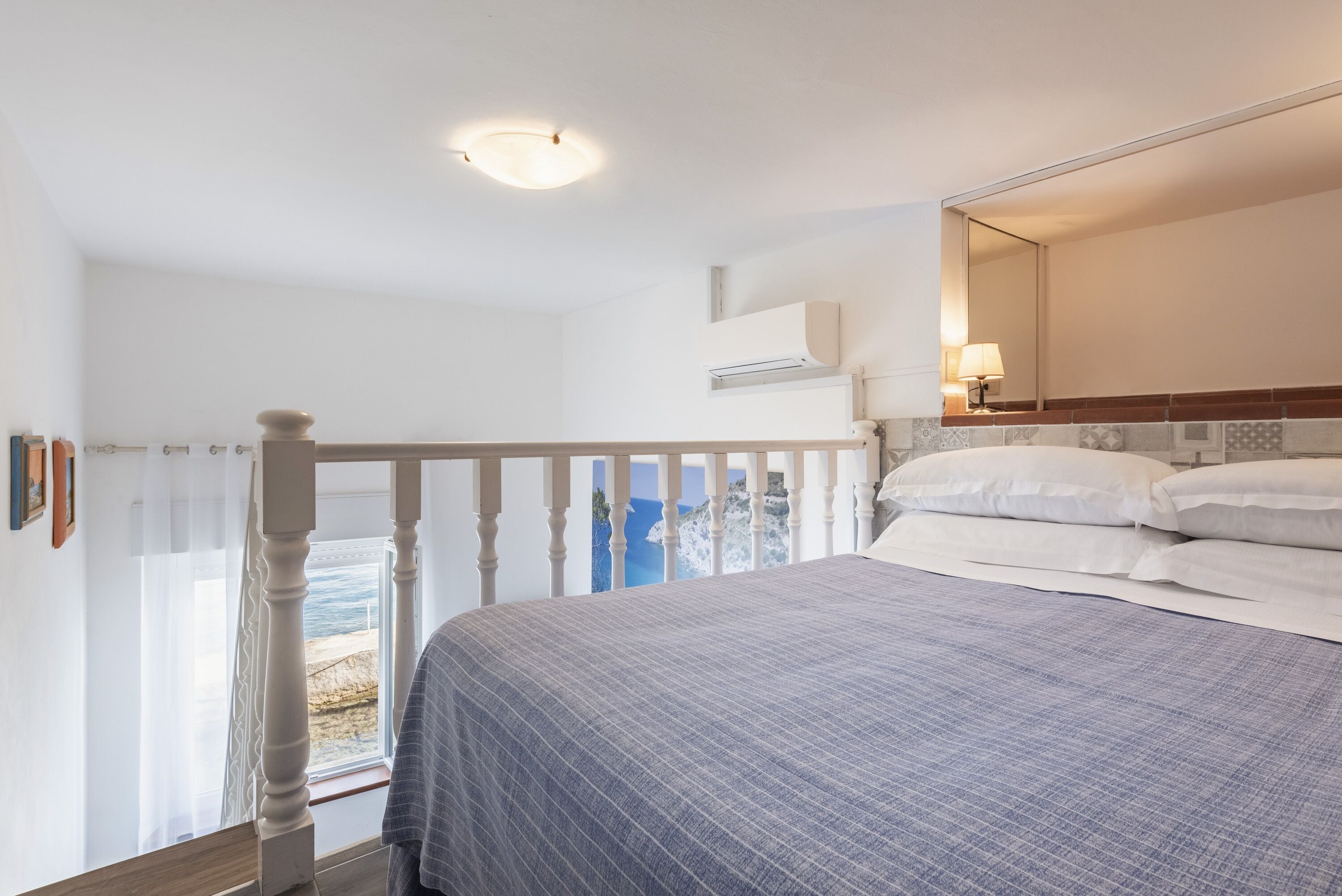 A bedroom with a bed, a window with a view of the sea, a ceiling light, and a wall-mounted air conditioner.