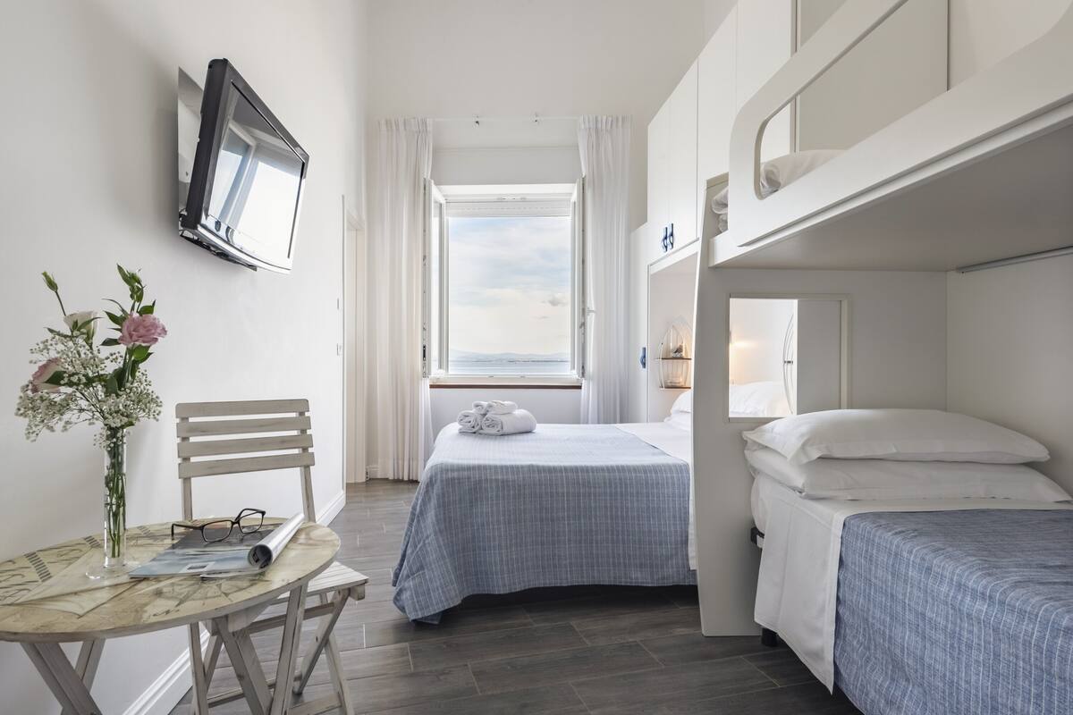 A compact hotel room with a bunk bed, a single bed, a small table with a vase of flowers, and a TV mounted on the wall.