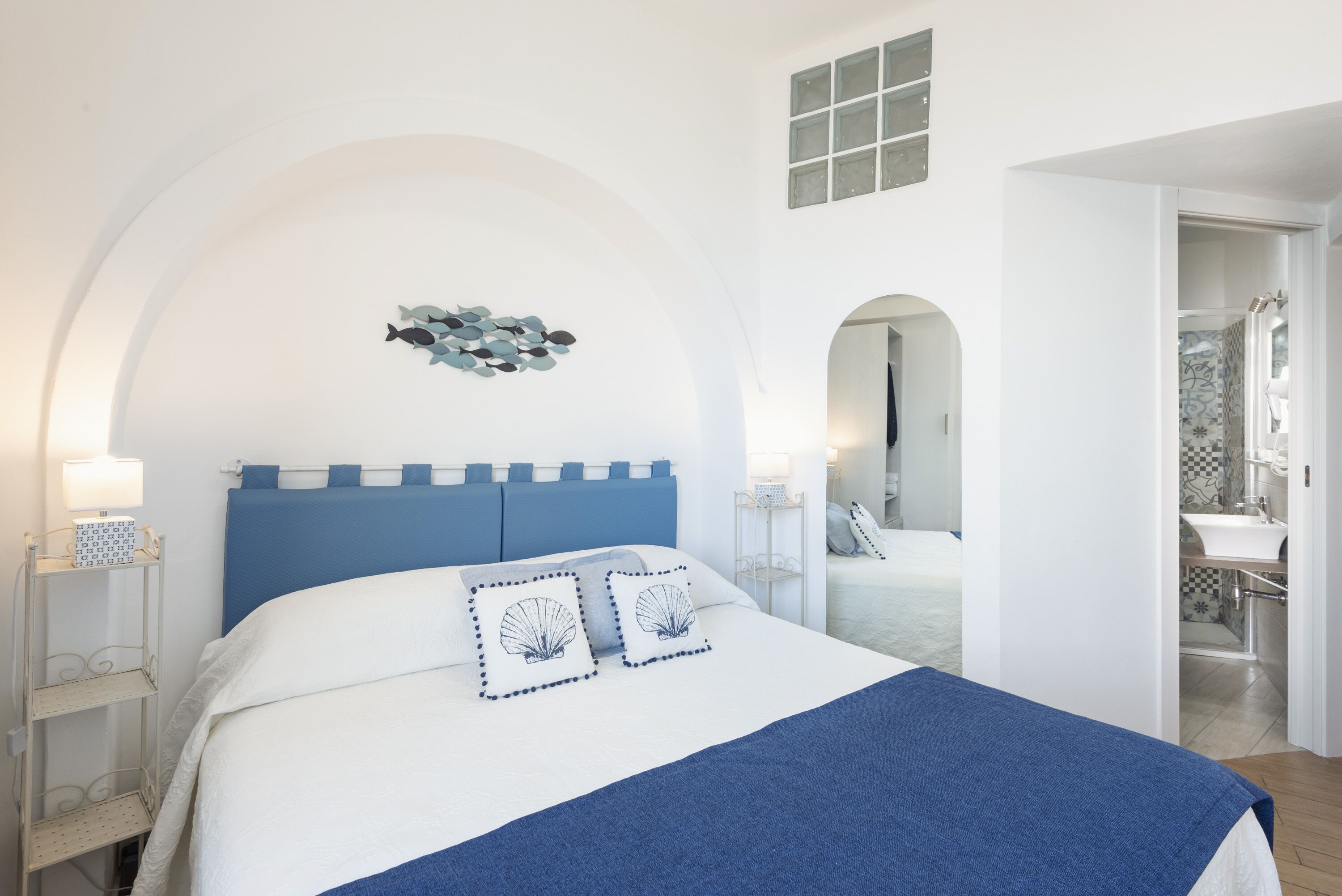 A bedroom with a bed, a nightstand, a wall-mounted lamp, and a view of a bathroom.