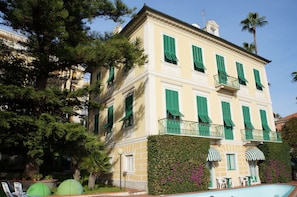 A two-story building with green shutters, a balcony, and a pool area.