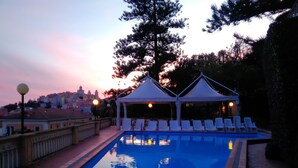 Una piscina con sillones de descanso y una zona de estar cubierta, rodeada de árboles y edificios al fondo.