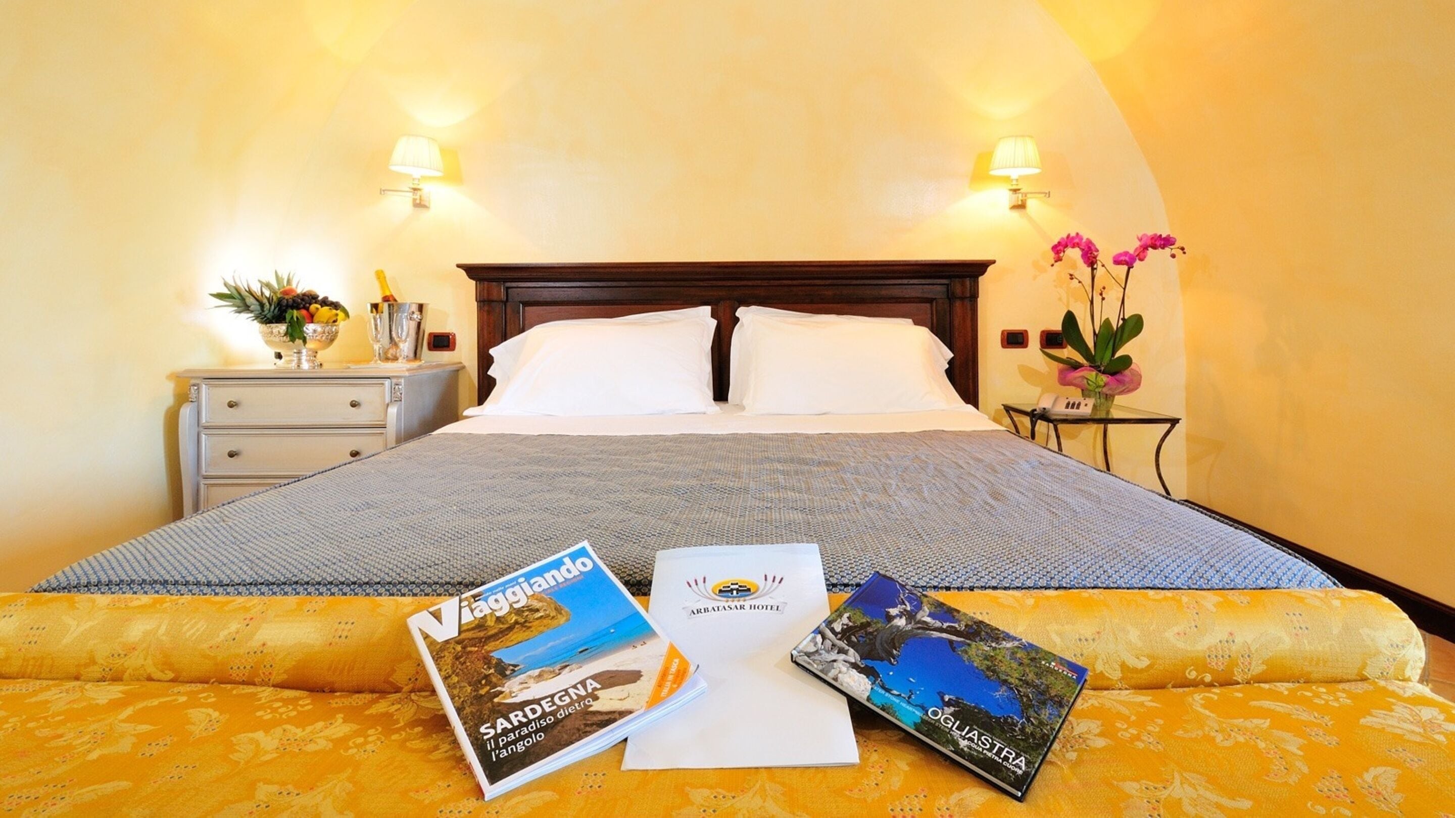 Camera d'albergo con un letto grande, due abat-jour, un comodino con lampada, un tavolino con un vaso di fiori e brochure sul letto.