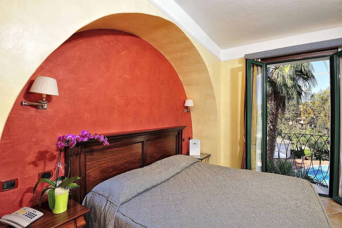 A hotel room with a large bed, a wooden headboard, a potted plant, a bedside table, and a view of a pool and greenery.