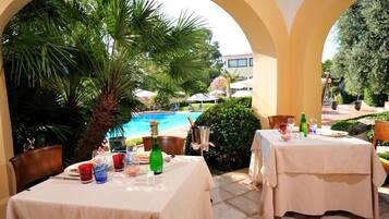 An outdoor dining area with tables set for two, overlooking a swimming pool and lush greenery.