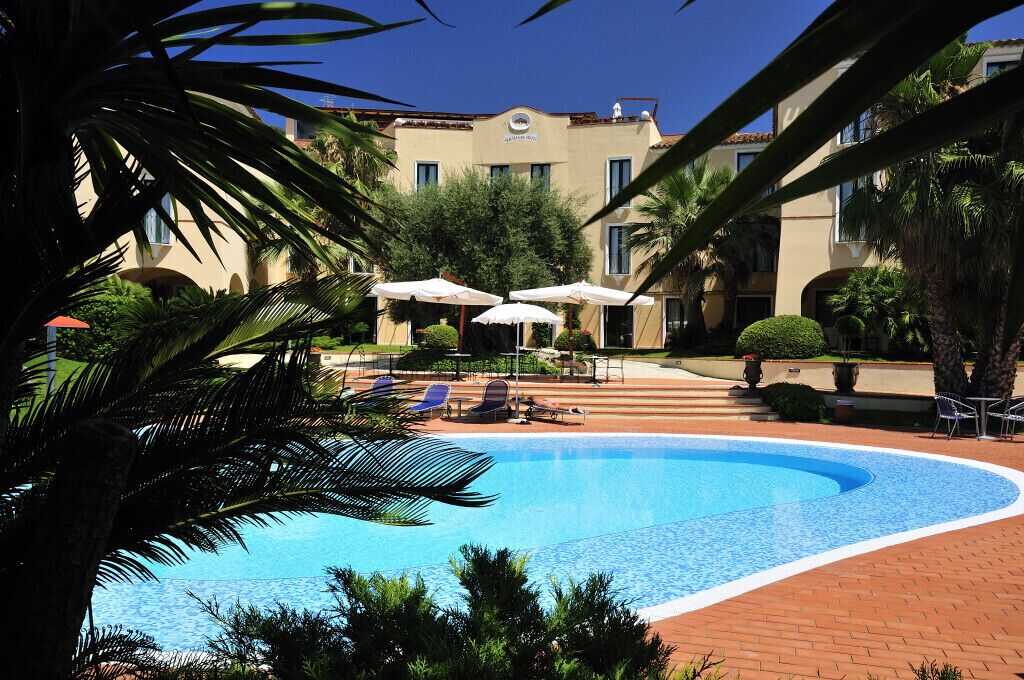 A swimming pool with lounge chairs and umbrellas, surrounded by palm trees and a building with a beige facade.