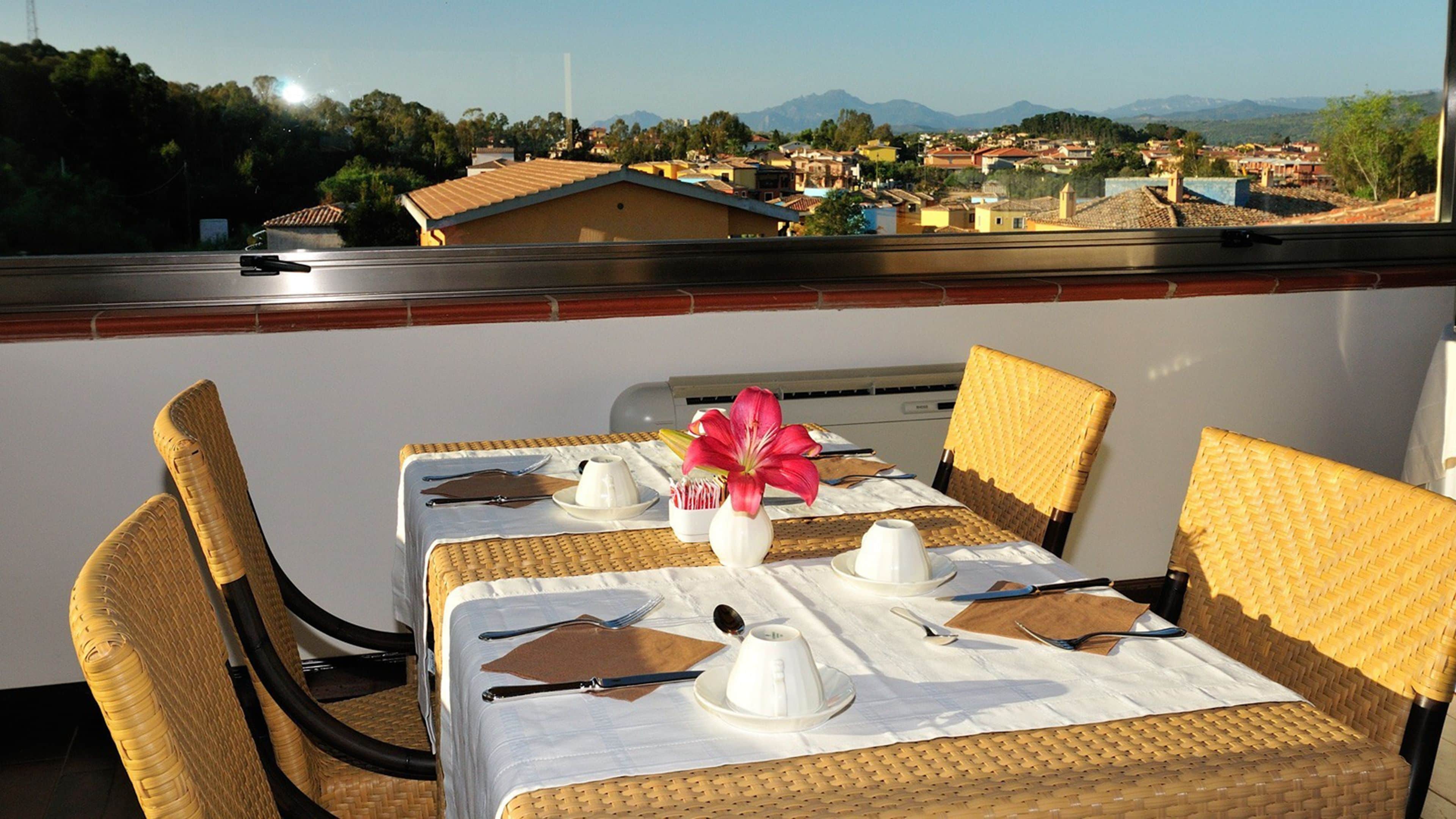 Una mesa de comedor dispuesta con vajilla blanca y un florero, con vistas a una zona residencial con casas y montañas.