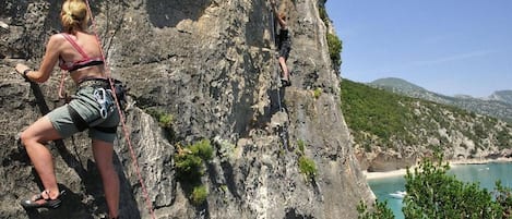 Dos personas escalan una pared rocosa con una playa y montañas al fondo.
