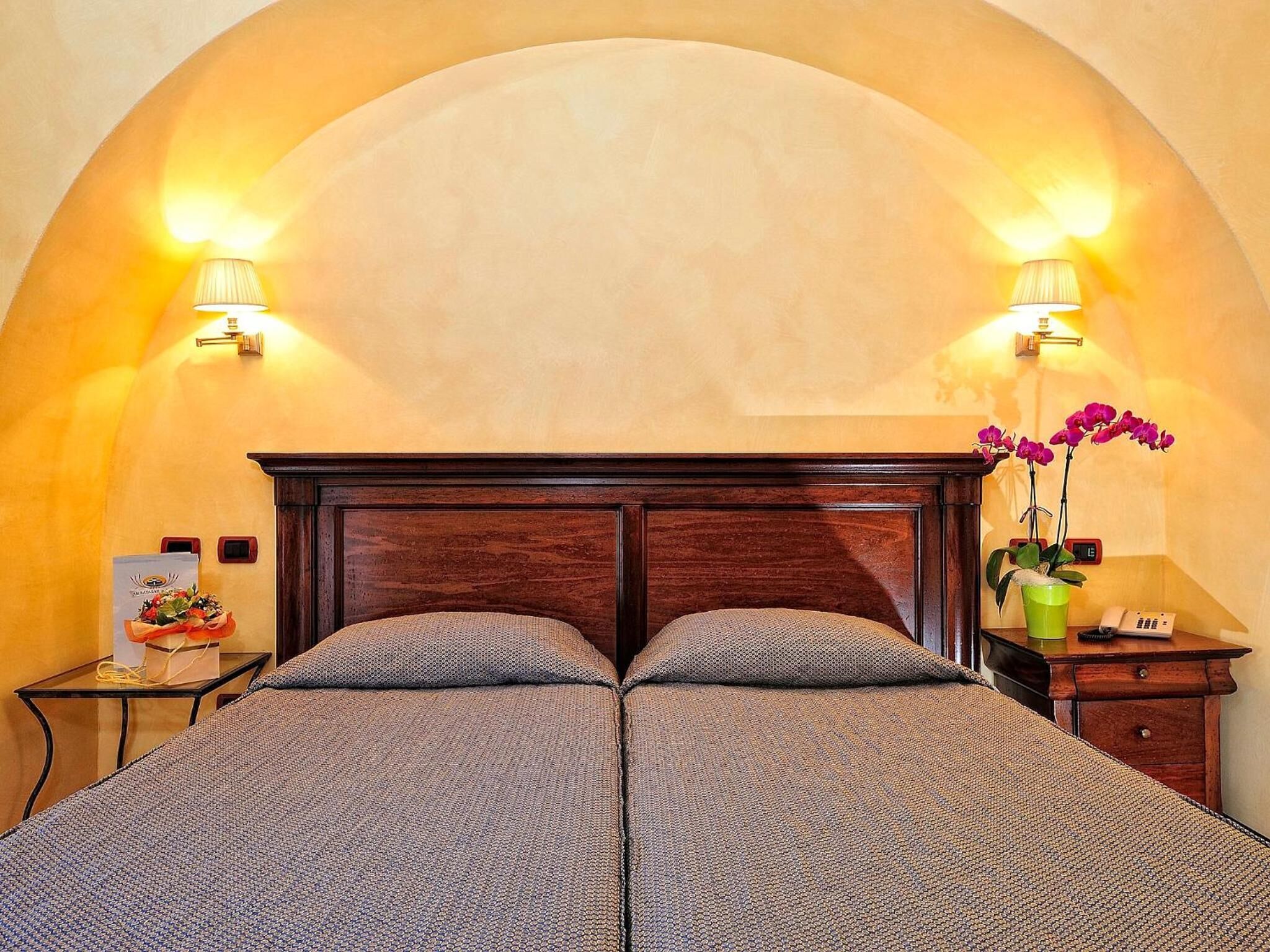 Una cama doble con cabecera de madera, dos lámparas de pared, una mesita de noche con un teléfono y un jarrón verde con flores, y una pequeña mesa con una cesta de fruta.