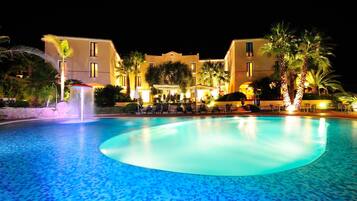 Una piscina iluminada con fuente, rodeada de palmeras y un edificio con múltiples ventanas y un letrero.