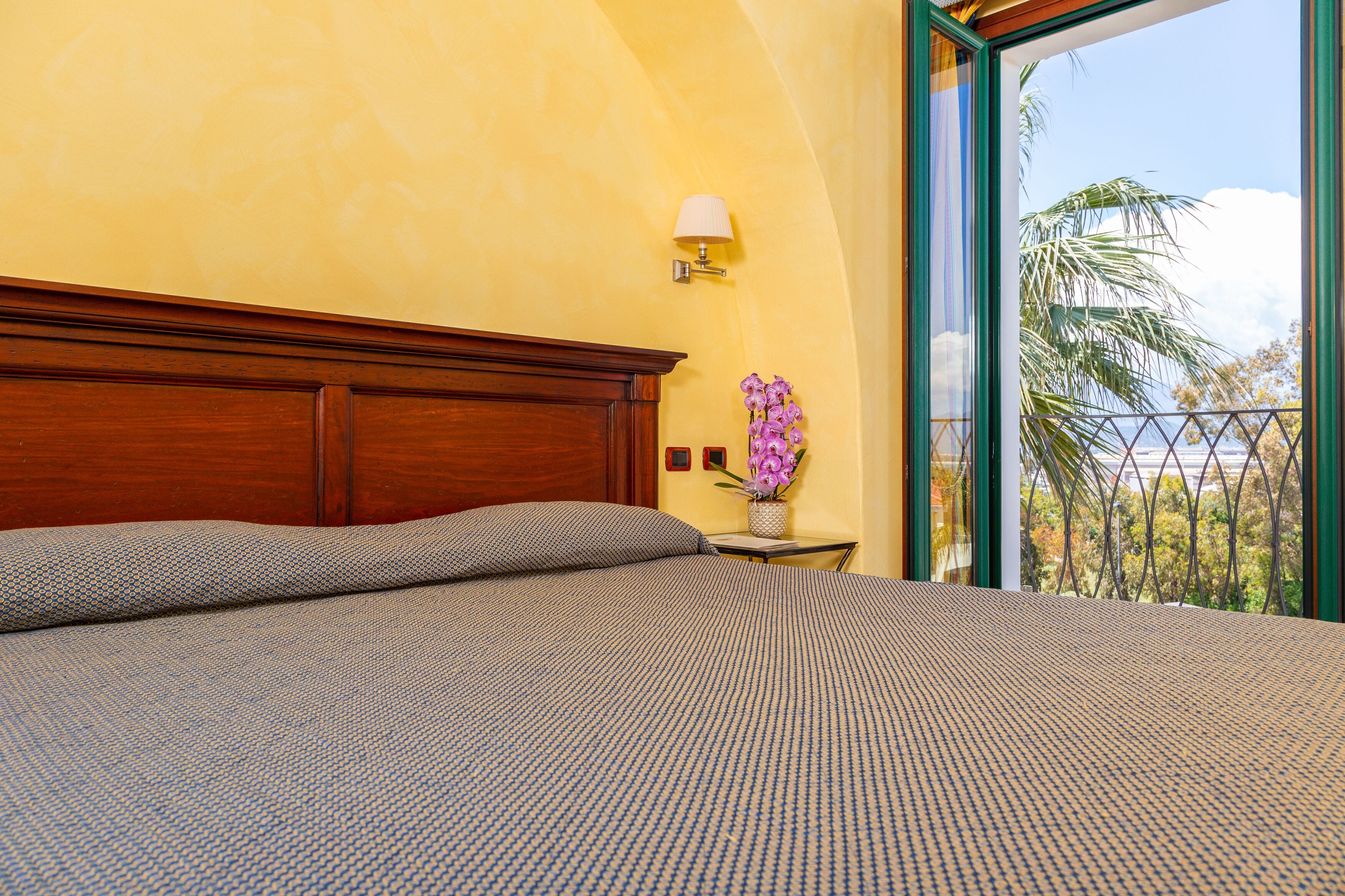 A bedroom with a large bed, a nightstand with a lamp, and a view of palm trees through a balcony.