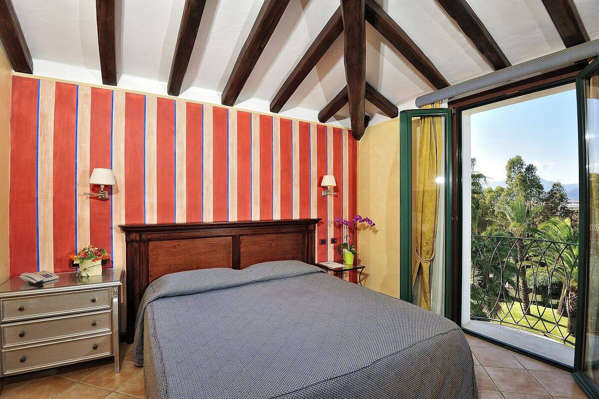 A bedroom with a wooden headboard, striped wallpaper, a bed with a grey bedspread, a nightstand with a lamp, a dresser, and a window with a view of greenery.