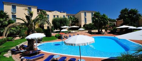 A pool area with lounge chairs and umbrellas, surrounded by palm trees and a building with multiple windows.