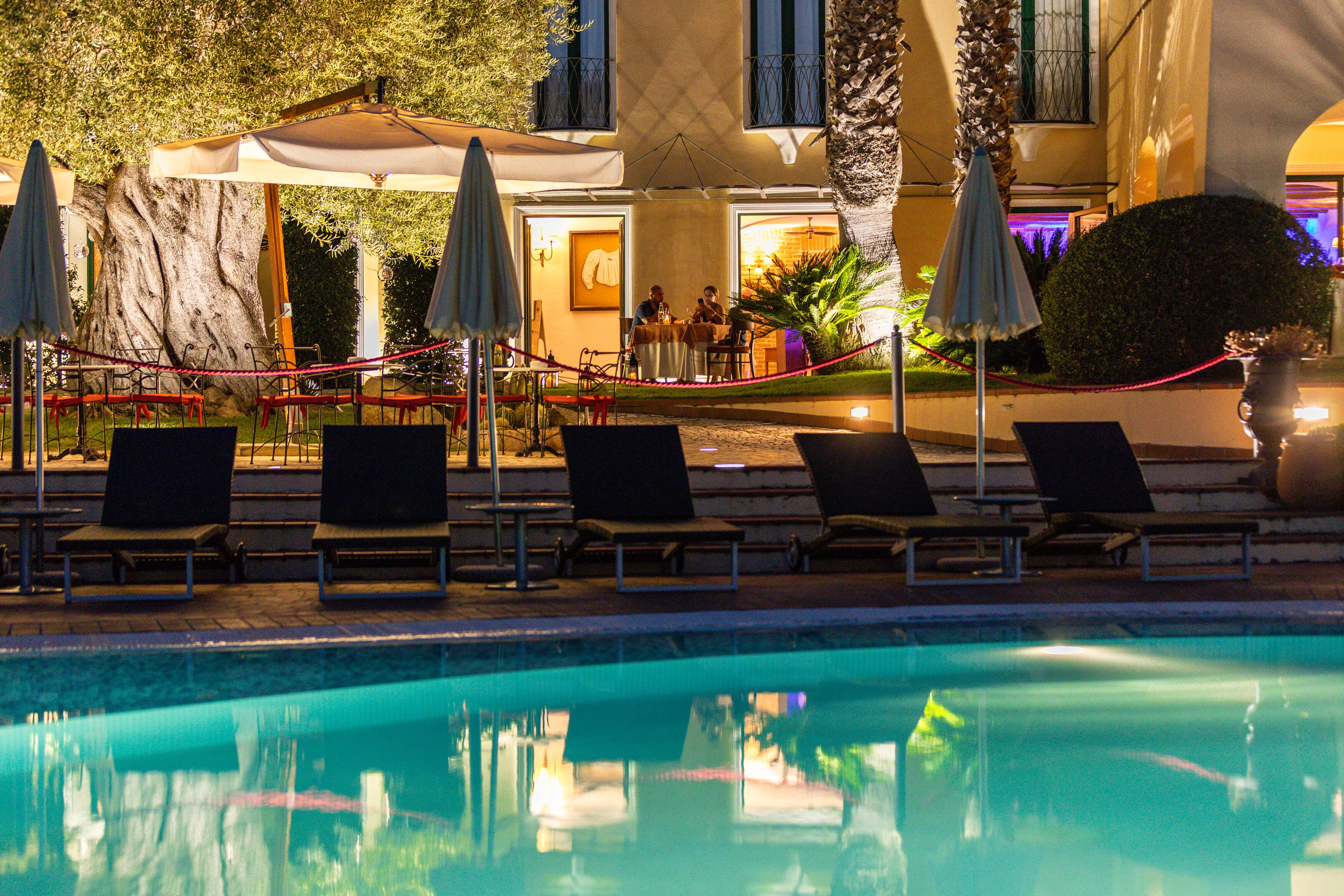 A poolside area with lounge chairs and umbrellas, a dining area with people, and a large tree.