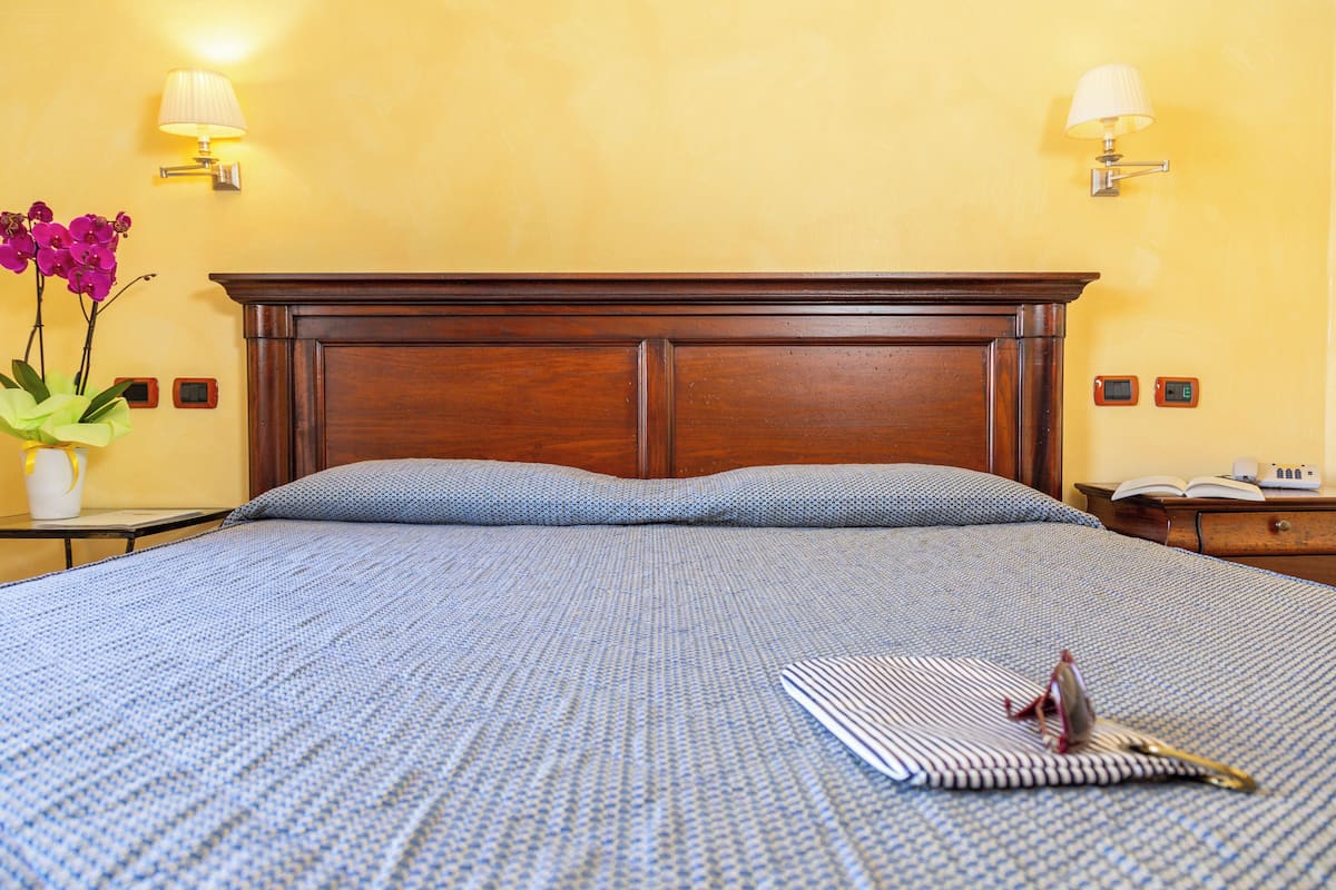 A neatly made bed with a blue patterned bedspread, a wooden headboard, and a bedside table with a lamp and a vase of flowers.