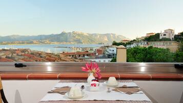 A table set with plates, cutlery, and a vase with a pink flower, overlooking a coastal town with buildings and mountains in the background.