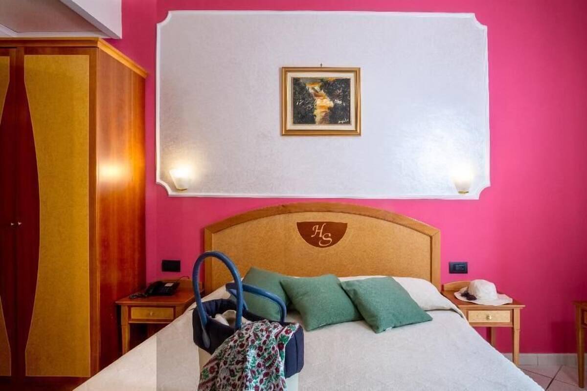 A hotel room with a bed, a headboard with a monogram, a bedside table, a nightstand, a wardrobe, and a wall-mounted picture.