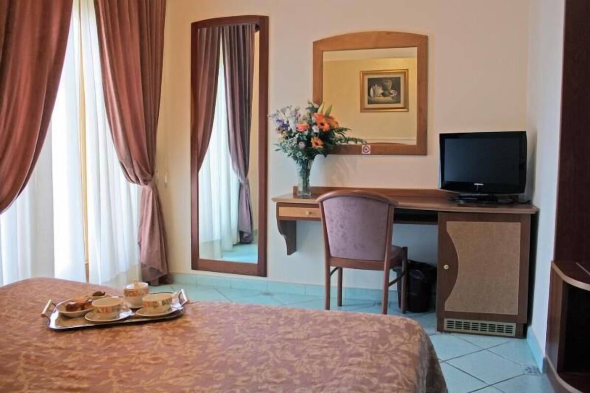 A hotel room with a bed, a desk with a television, a chair, and a tray with breakfast items.
