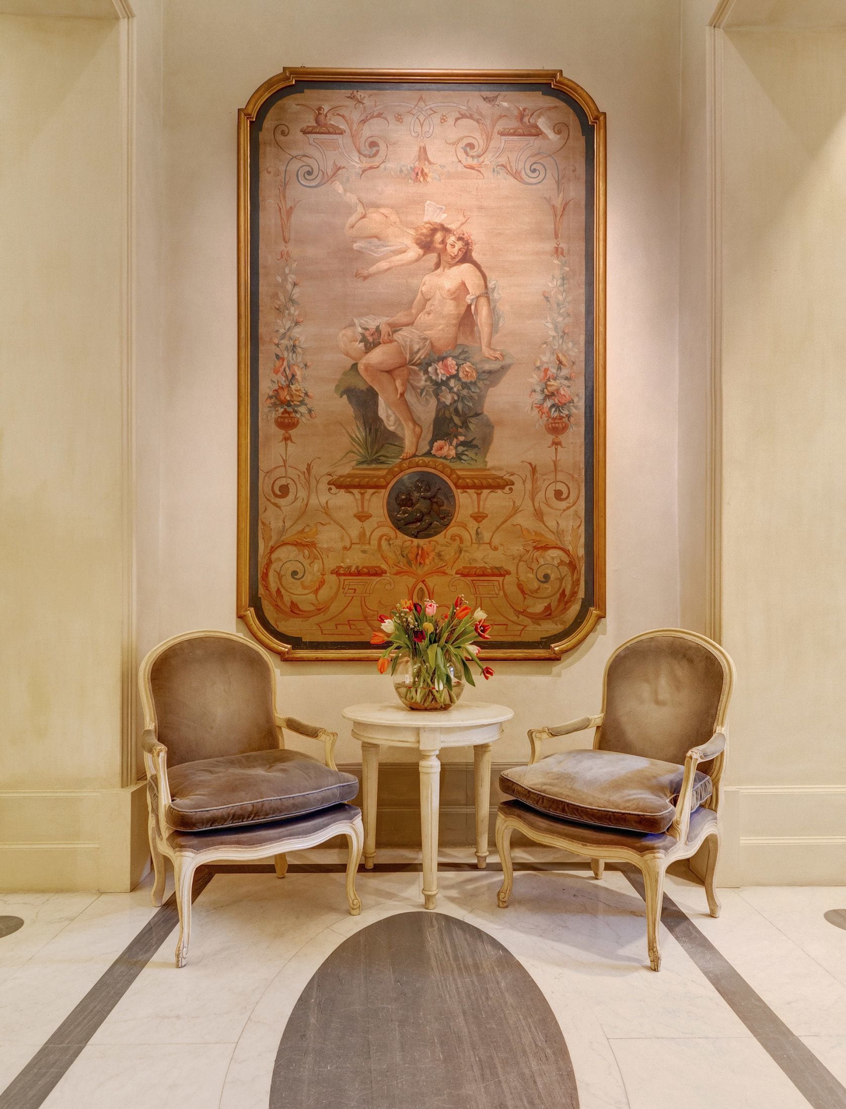 Una stanza con due poltrone imbottite, un tavolino con un vaso di fiori e un grande quadro decorativo appeso al muro.