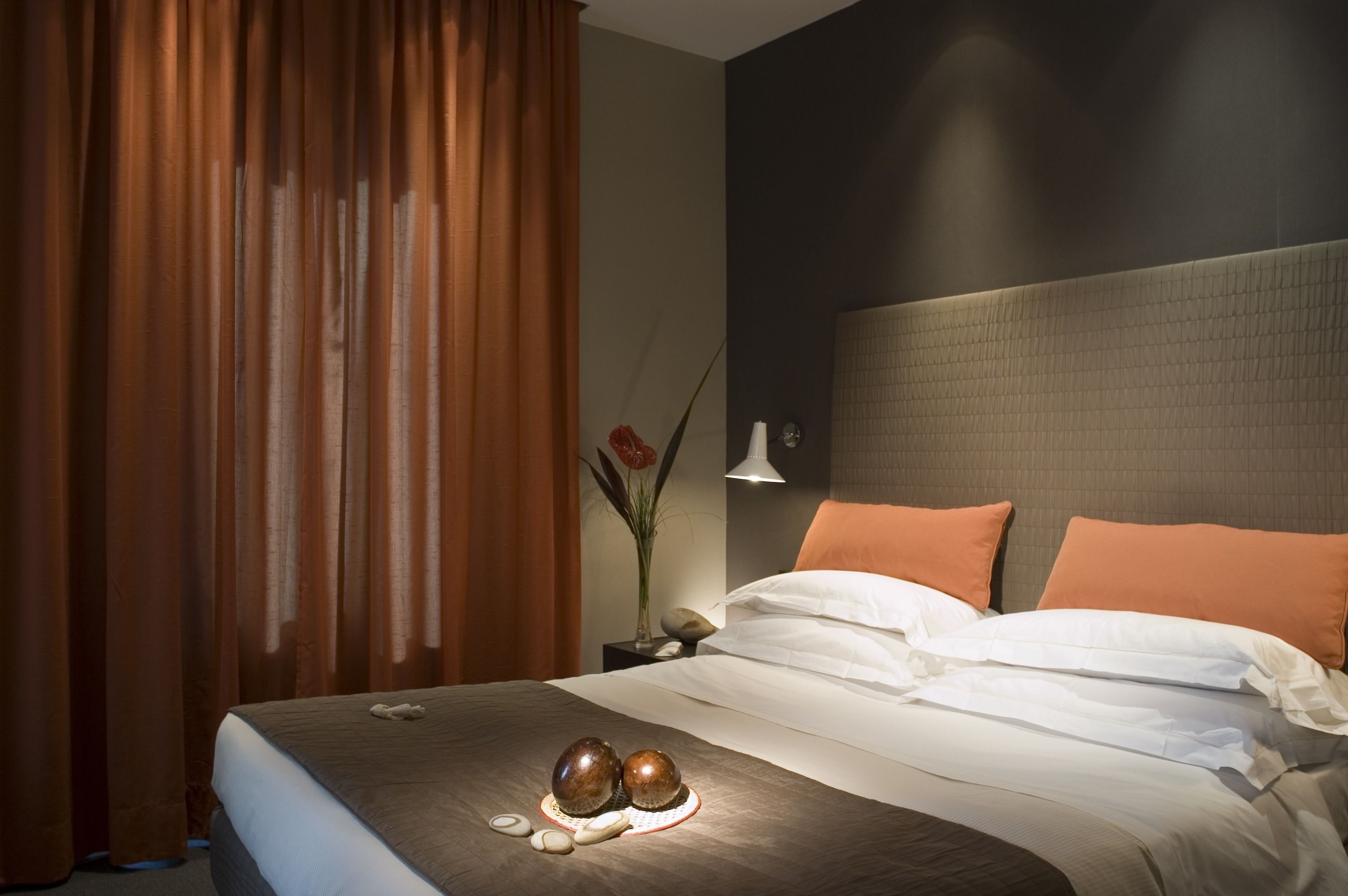 A hotel room with a bed, brown curtains, a vase with flowers, and a nightstand with a lamp.