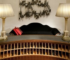 Una stanza con un divano nero, un cuscino rosso, due lampade e un'opera d'arte a parete.