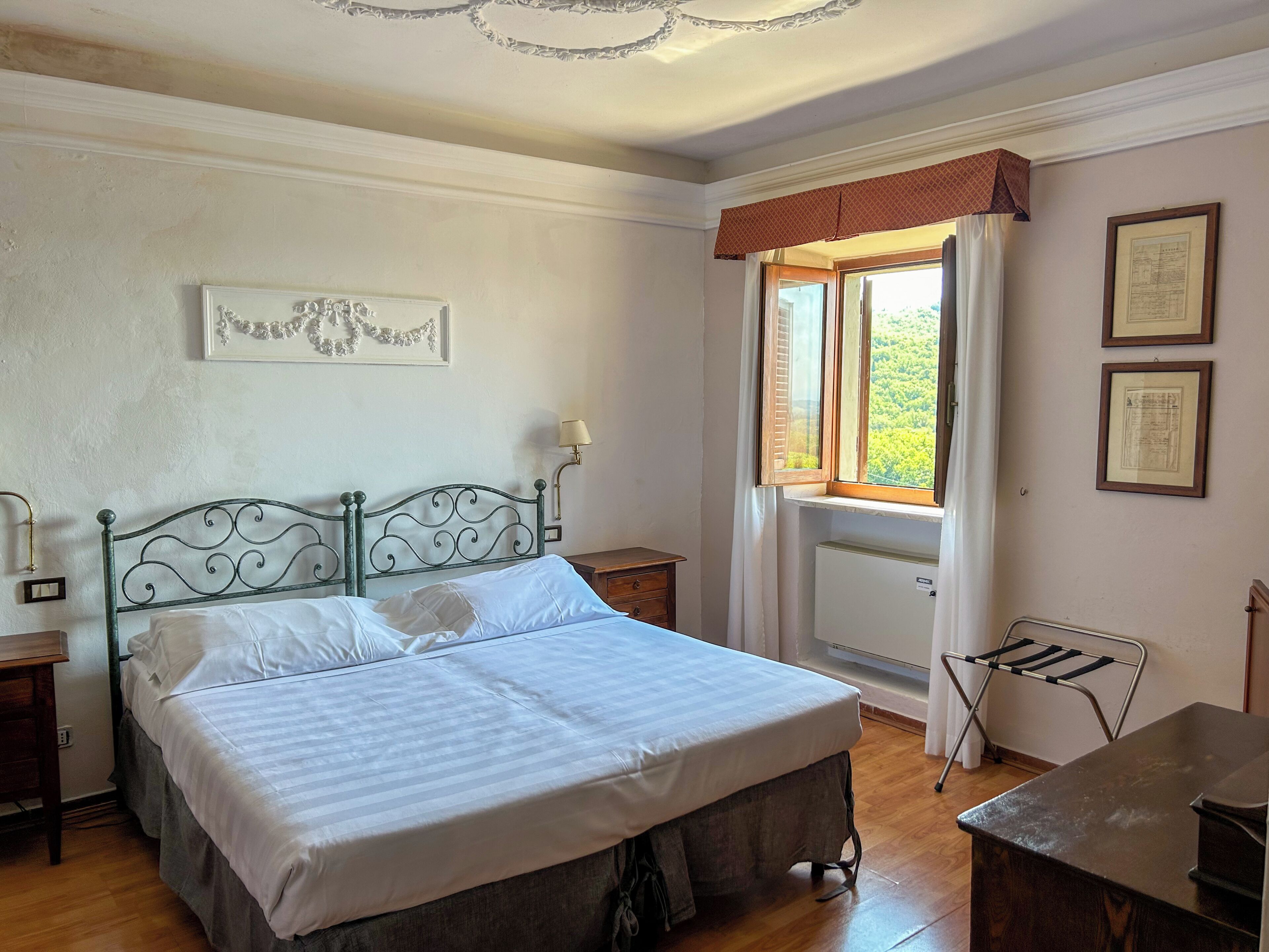 A bedroom with a bed, a nightstand, a lamp, a window with curtains, and a radiator.