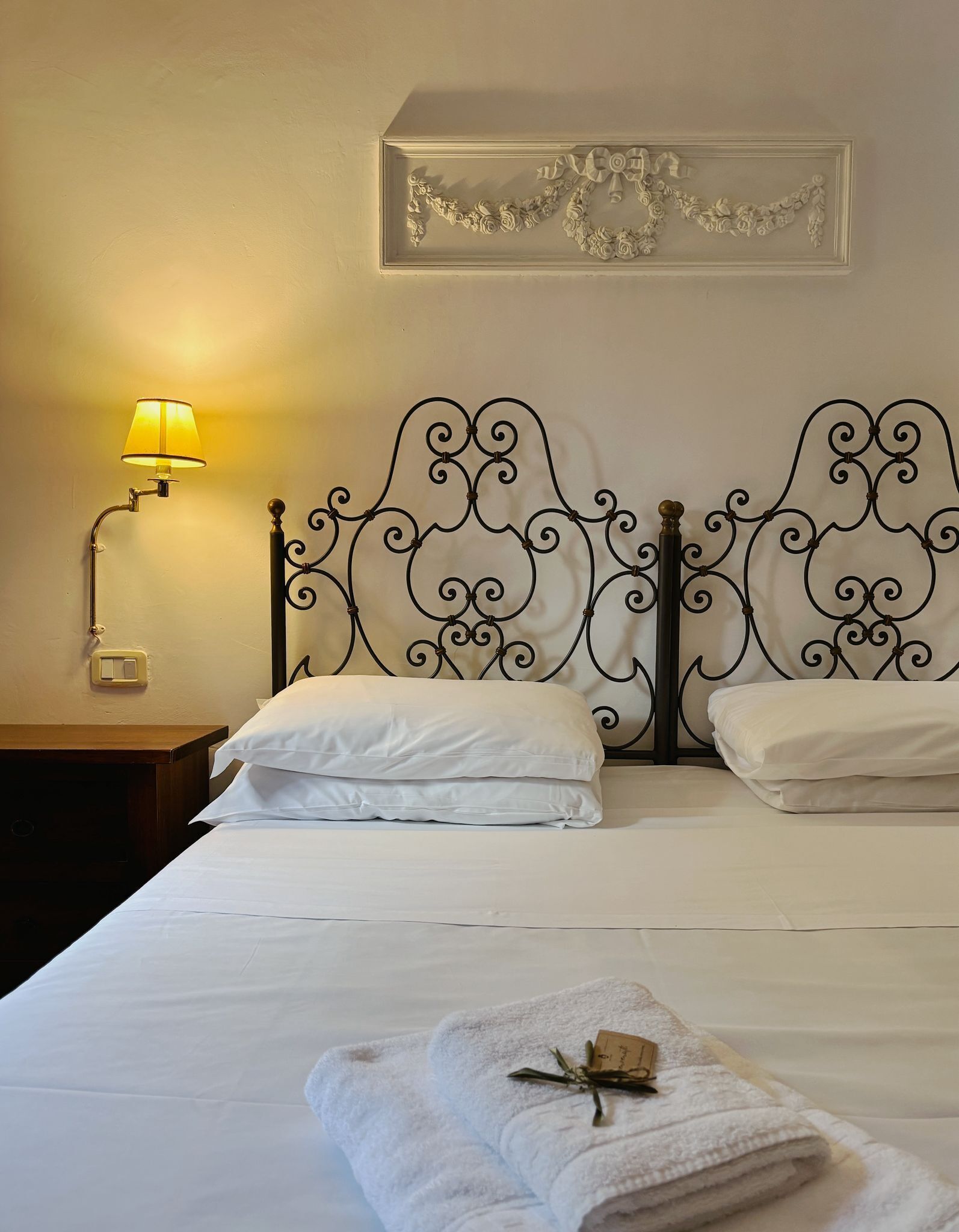 Una cama bien hecha con sábanas blancas, cabecera de metal con detalles ornamentales y un adorno en la pared, además de una lámpara de noche.