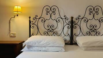 Una cama bien hecha con sábanas blancas, cabecera de metal con detalles ornamentales y un adorno en la pared, además de una lámpara de noche.