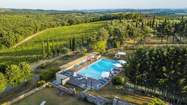 An aerial view of a villa with a swimming pool, surrounded by lush greenery and a hilly landscape.