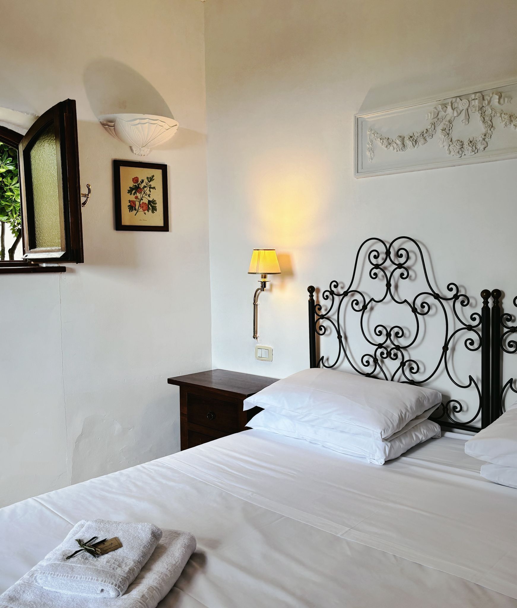 A neatly made bed with white linens, a bedside lamp, a framed botanical print, and a decorative wall piece.
