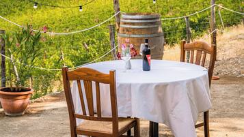 Uma área de refeições ao ar livre com mesa e cadeiras, um barril de vinho e vinhedos ao fundo.