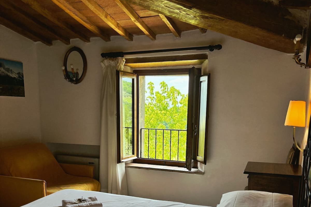 A bedroom with a bed, a window with a view of trees, a lamp, and a wooden ceiling.